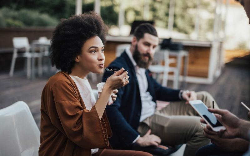 woman sits and eats outside while someone holding a cell talks to her